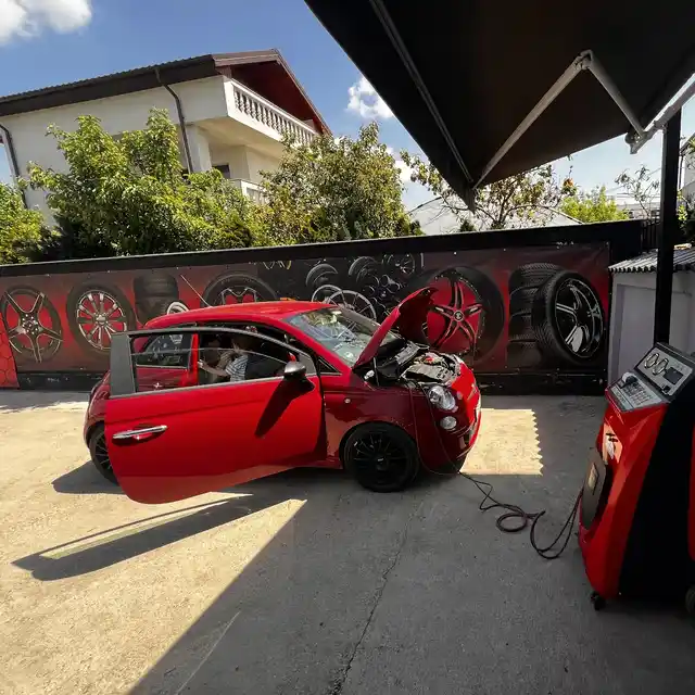 Un Fiat 500 roșu cu capota ridicată, legat la aparatul de freon, în curte.